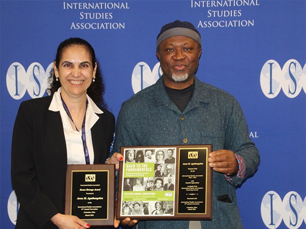 Two individuals stand in front of a backdrop with "International Studies Association" and its acronym, "ISA," prominently displayed. Each person holds a plaque featuring the ISA logo.