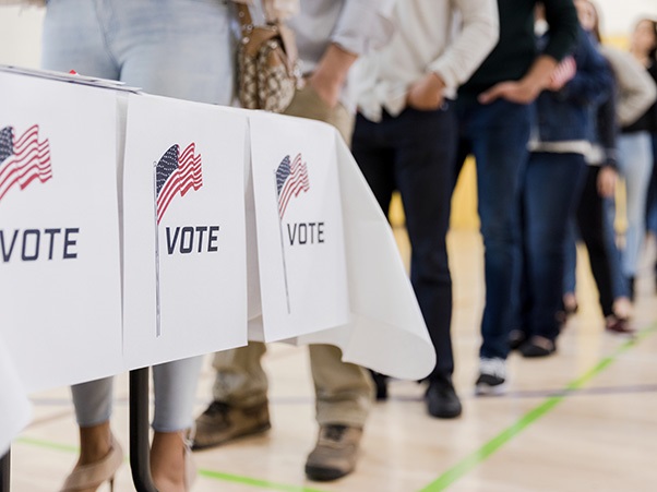 People wait in line at a polling place beside a table with “Vote” signs bearing American flags.