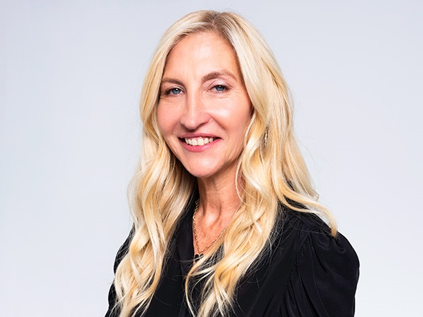 A person with long blonde hair, wearing a black top, smiling against a light gray background.