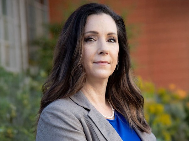 A person with long dark brown wavy hair, wearing a gray blazer over a blue top and a pearl necklace, photographed outdoors against a blurred background of greenery and a brick building.
