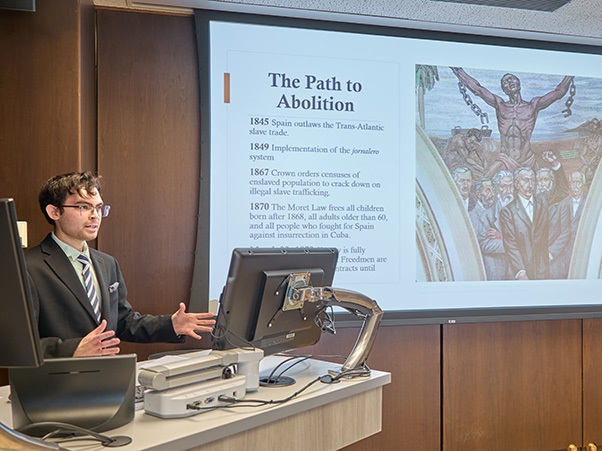 A person is giving a presentation in a lecture room. The slide on the screen is titled "The Path to Abolition" with historical dates and events listed, accompanied by a statue image. The room includes a podium and a computer setup.