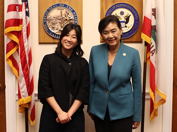 Two people smiling, flanked by an American flag and a California state flag. Behind them are two official seals mounted on the wall, including the Great Seal of the State of California and a U.S. House of Representatives seal.