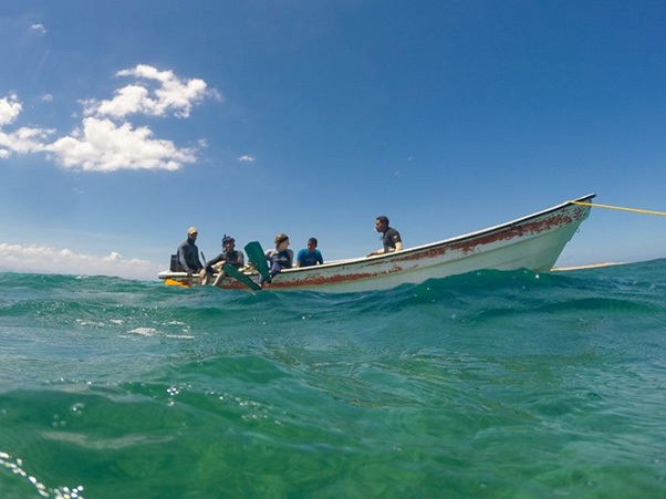 A small group of people sits in a boat on clear, turquoise water under a bright blue sky with scattered clouds.