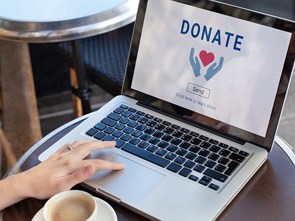A person is sitting at a round table with a coffee cup, using a laptop. The laptop screen displays the word "DONATE" with an icon of hands holding a heart, and a button labeled "Send.