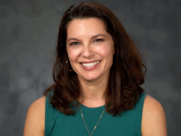 A person smiling with shoulder-length dark hair, wearing a teal sleeveless top with a thin gold necklace and gold hoop earring, against a mottled gray backdrop.