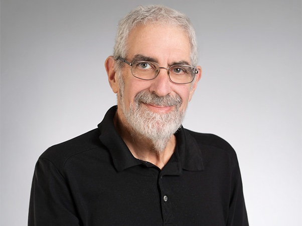 A smiling person with short gray hair, a gray beard, and glasses, wearing a black collared polo shirt against a light gray background.
