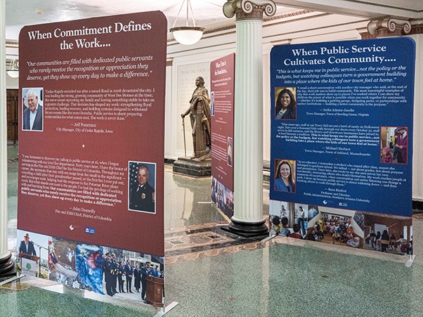 Exhibit banners on public service displayed among columns near a statue.
