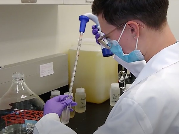 Researcher wearing lab coat, mask and gloves uses a pipette on wastewater sample bottles.