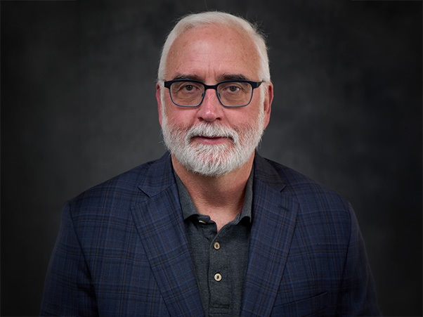 A person with white hair, a white beard, and glasses, wearing a dark plaid blazer over a gray polo shirt, set against a dark gray background.