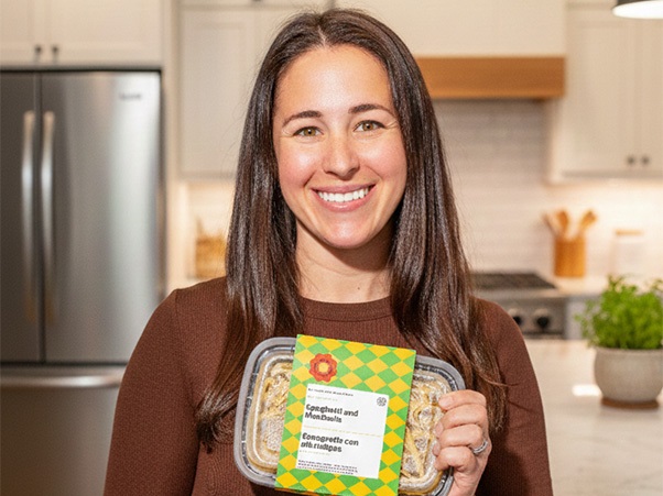 A person smiles and holds a packaged prepared meal in a kitchen setting.