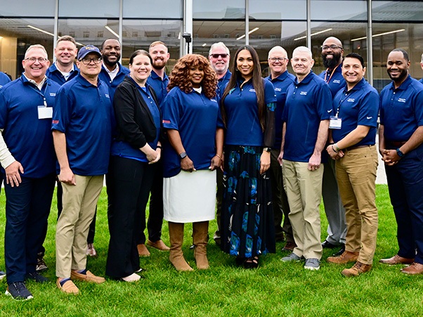 A group of thirteen people stands on a grassy area in front of a modern building with large windows. They are all wearing matching blue polo shirts. The individuals are posing and smiling.