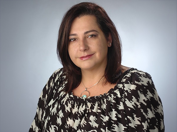 A person wearing a black-and-white patterned top and a delicate necklace with a turquoise pendant, against a neutral gray background.