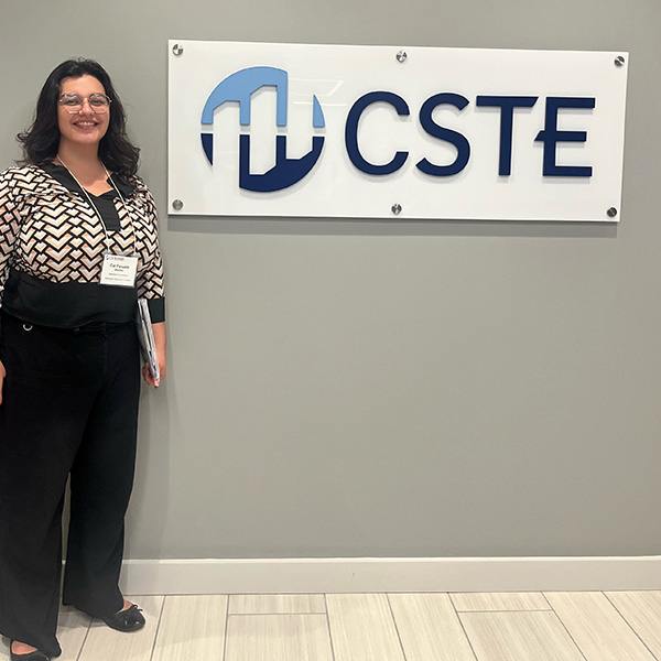 A person stands next to a wall-mounted CSTE sign, smiling at the camera. She has long dark wavy hair, wears glasses, and is dressed in a black-and-white patterned blouse and black pants.