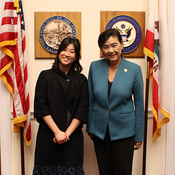 Two individuals stand smiling in an office setting between the flags of the United States and California. Behind them are plaques with official seals.